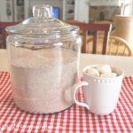 A cup of coffee with a few marshmallows on top, placed beside a jar of homemade hot chocolate mix, sits on a table.