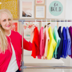 woman with blonde hair smiling at the camera and standing next to a rack of colorful cardigans that have been hang folded