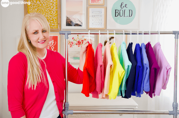woman with blonde hair smiling at the camera and standing next to a rack of colorful cardigans that have been hang folded