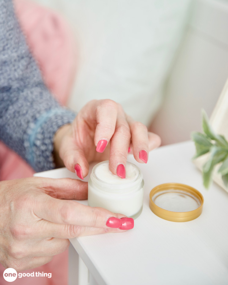 A woman is putting cream into a jar on a table to relieve her everyday aches.