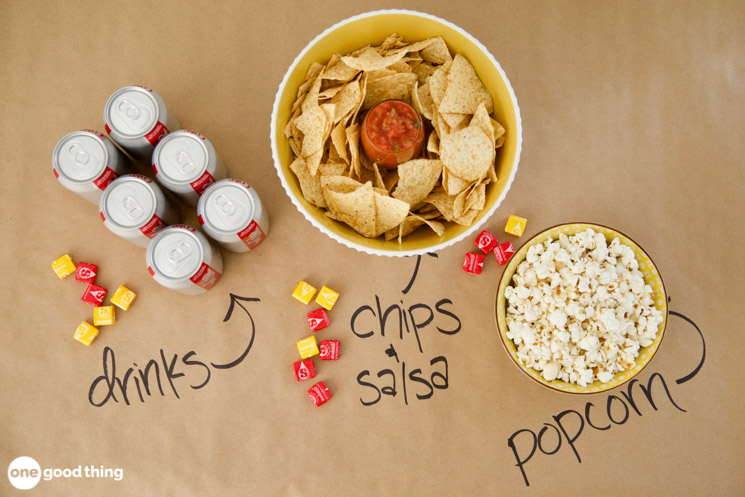 table covered in kraft paper with diet coke, chips and salsa, and popcorn sitting on the table and the labels for those three things have been written on the kraft paper in sharpie