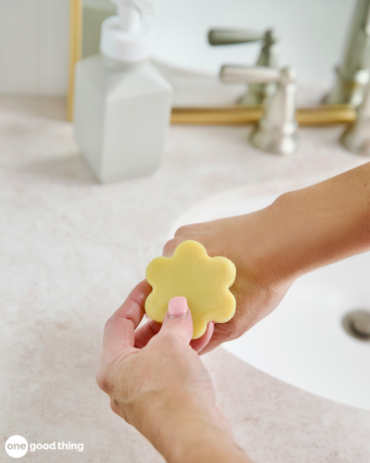 A person holding a yellow flower soap, known for its moisturizing properties, in front of a sink.