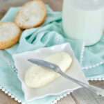 making butter and buttermilk - homemade butter and butter knife on a small square plate, a cut biscuit, and a bottle of buttermilk, all on a light aqua cloth