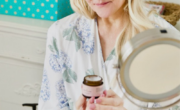 A woman examines a cosmetic product for aging skin while sitting at a table with various skincare products and a magnifying mirror.