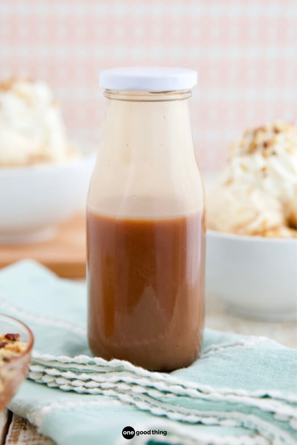 A glass bottle filled with rich brown liquid, topped with a white cap, placed on a light blue cloth with bowls of hot fudge dessert in the background.