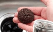 A hand holding a ball of used coffee grounds near a kitchen sink, suggesting using old coffee grounds for cleaning purposes. Text reads: "don't throw your old coffee grounds away! TRY THIS CLEANING HACK FOR YOUR GARBAGE DISPOSAL.