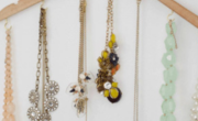 A wooden hanger, adorned with assorted necklacesโbeaded, chain, and charm stylesโgraces a white wall.