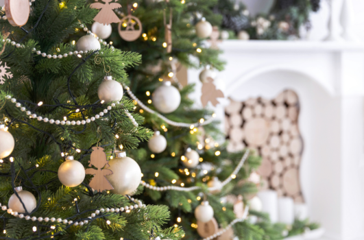 A decorated Christmas tree with white and gold ornaments and lights graces the room, capturing this year's top holiday decorating ideas. In the background, a white fireplace filled with stacked logs completes the cozy and elegant atmosphere.