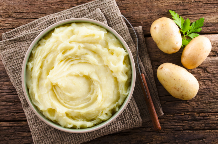A bowl of creamy mashed potatoes rests on a wooden surface, surrounded by three whole potatoes and a sprig of parsley, offering tasty ways to celebrate Mashed Potato Day.