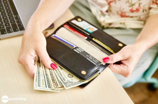 A person holding an open wallet with cash and credit cards next to a laptop on a desk contemplates the budgeting rule.