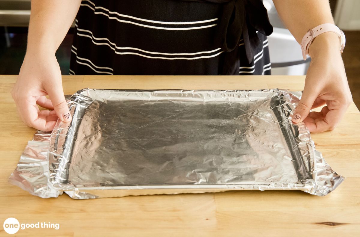 dishwashing hacks_5 A person covers a baking sheet with aluminum foil on a wooden table, preparing for easy dishwashing afterward.