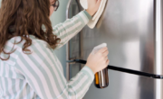 A woman cleans stainless steel appliances, wiping down a refrigerator with a cloth in one hand and a spray bottle in the other.