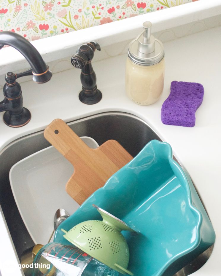 dishwashing hacks_2 A kitchen sink with a wooden cutting board, teal dish rack, colander, glass, soap dispenser, and purple sponge on a white counter—everything you need for easy dishwashing.