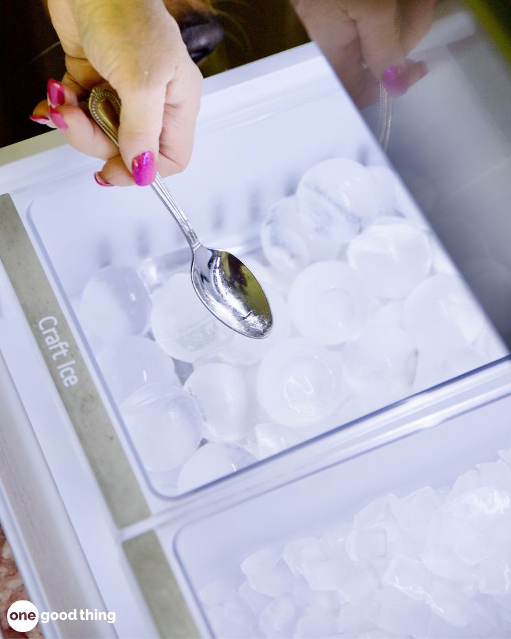 A person, trying to soothe mosquito bites, scoops craft ice cubes from a freezer drawer using a metal spoon.