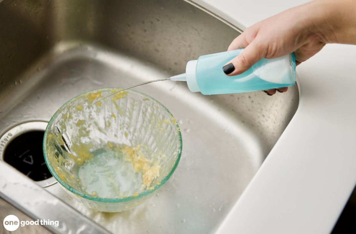 dishwashing hacks_6 A person squirts blue dish soap into a glass bowl with food residue in a stainless steel kitchen sink, getting ready for dishwashing.