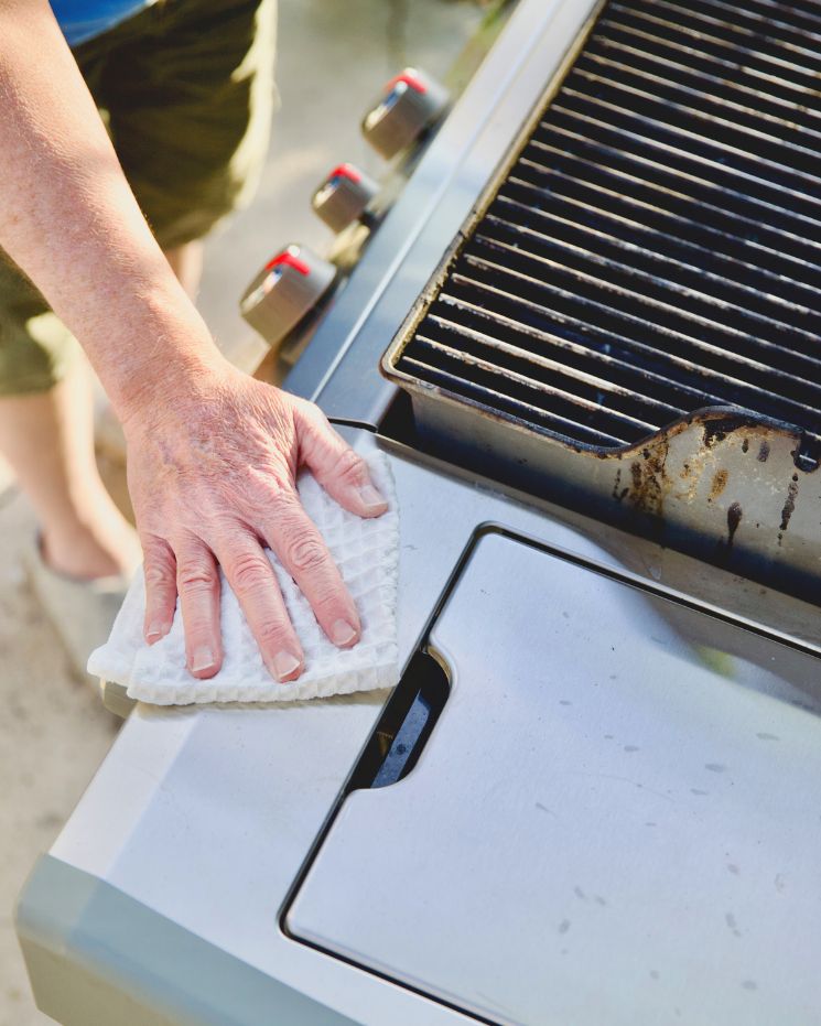 practical father's day gift_6 A person wipes the surface of an outdoor gas grill with a white cloth, cleaning near the grill’s controls and cooking area—showing why this makes such a practical gift or useful gift for Father’s Day.