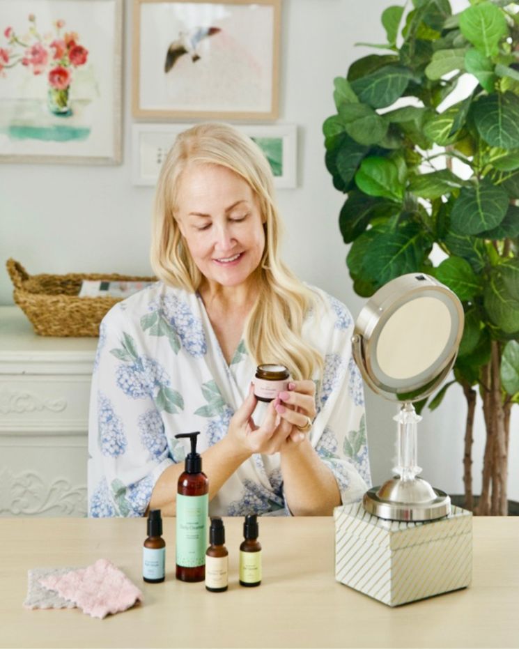skincare confessions_8 A woman in a floral robe sits at a table, holding a skincare product jar, as if ready to share her skincare confessions. Several other bottles are lined up in front of her next to a tabletop mirror.