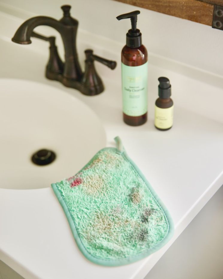 skincare confessions_7 A stained green washcloth lies on a white bathroom counter near a sink, next to two bottles of skincare products—an embarrassing skincare confession left in plain sight.