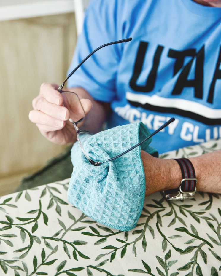 practical father's day gift_4 A person wearing a blue Utah Hockey Club shirt is cleaning eyeglasses with a blue cloth at a table with a leafy patterned tablecloth—a practical gift idea, perfect as a Father’s Day gift.