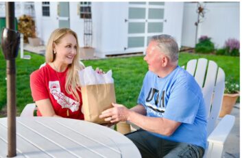 A woman hands a paper bag with tissue paper to an older man as they sit outside at a patio table in a backyard, sharing a special Father’s Day gift.