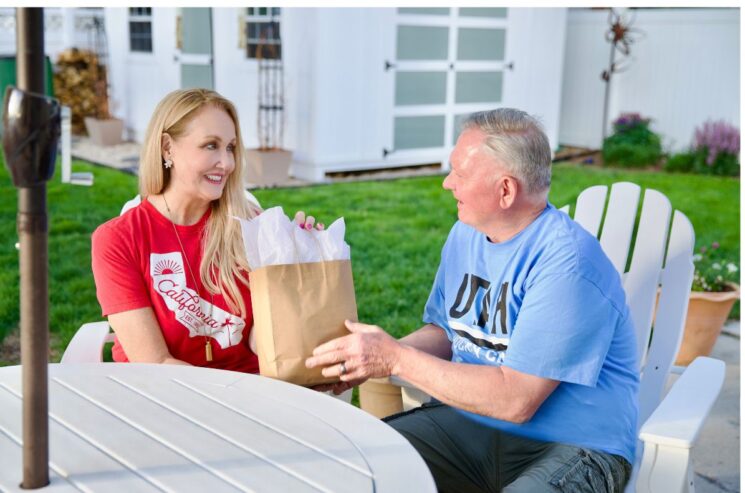 A woman hands a paper bag with tissue paper to an older man as they sit outside at a patio table in a backyard, sharing a special Father’s Day gift.
