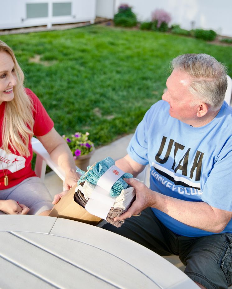 practical father's day gift_10 Two people sit at an outdoor table; the man hands a set of folded clothes—a practical Father’s Day gift—to the woman, who is smiling. Grass and a flower pot are visible in the background.