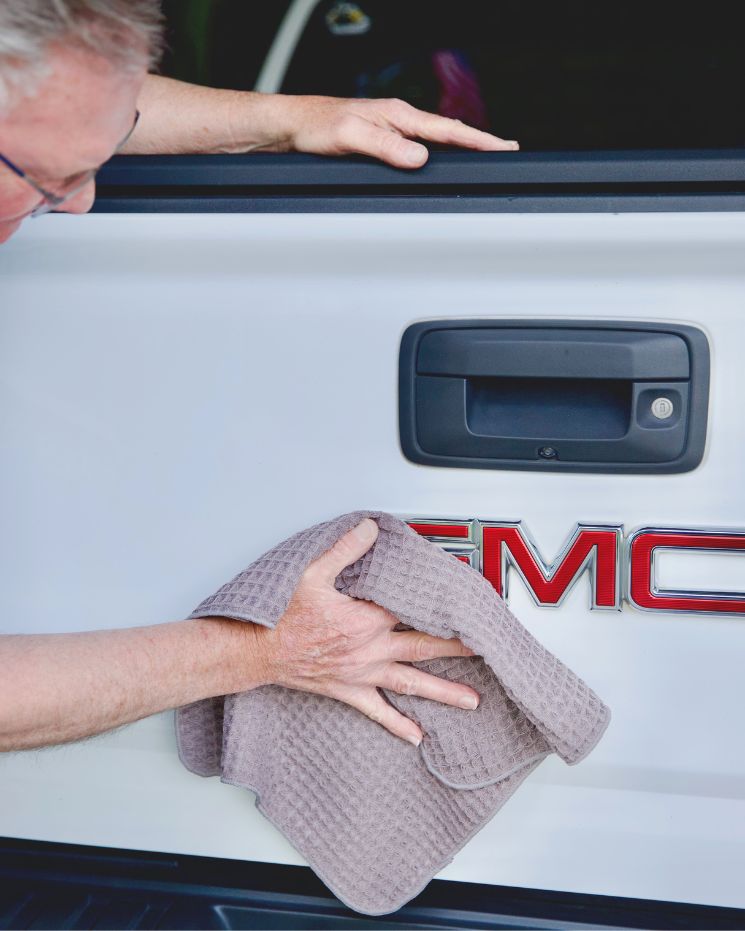 practical father's day gift_3 A person wipes the back of a white GMC vehicle with a beige cloth, focusing on the area near the logo and handle—a practical gift idea for Father's Day.