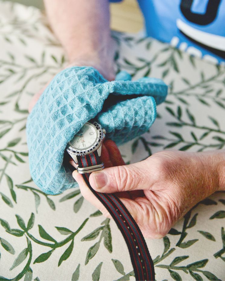 practical father's day gift_7 A person wipes a wristwatch with a blue textured cloth while holding it over a patterned tablecloth—a thoughtful and practical Father’s Day Gift in the making.