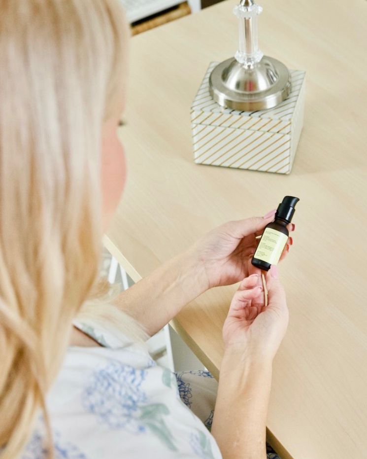 skincare confessions_5 A person with blonde hair sits at a desk, holding a small bottle with a pump dispenser and yellow label—perhaps ready for some skincare confessions—next to a lamp and a decorative box.