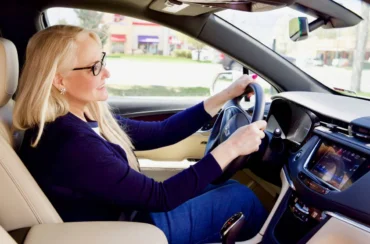 A woman with long blonde hair and glasses is driving a car, smiling and looking ahead, enjoying summer travel with her hands on the steering wheel.