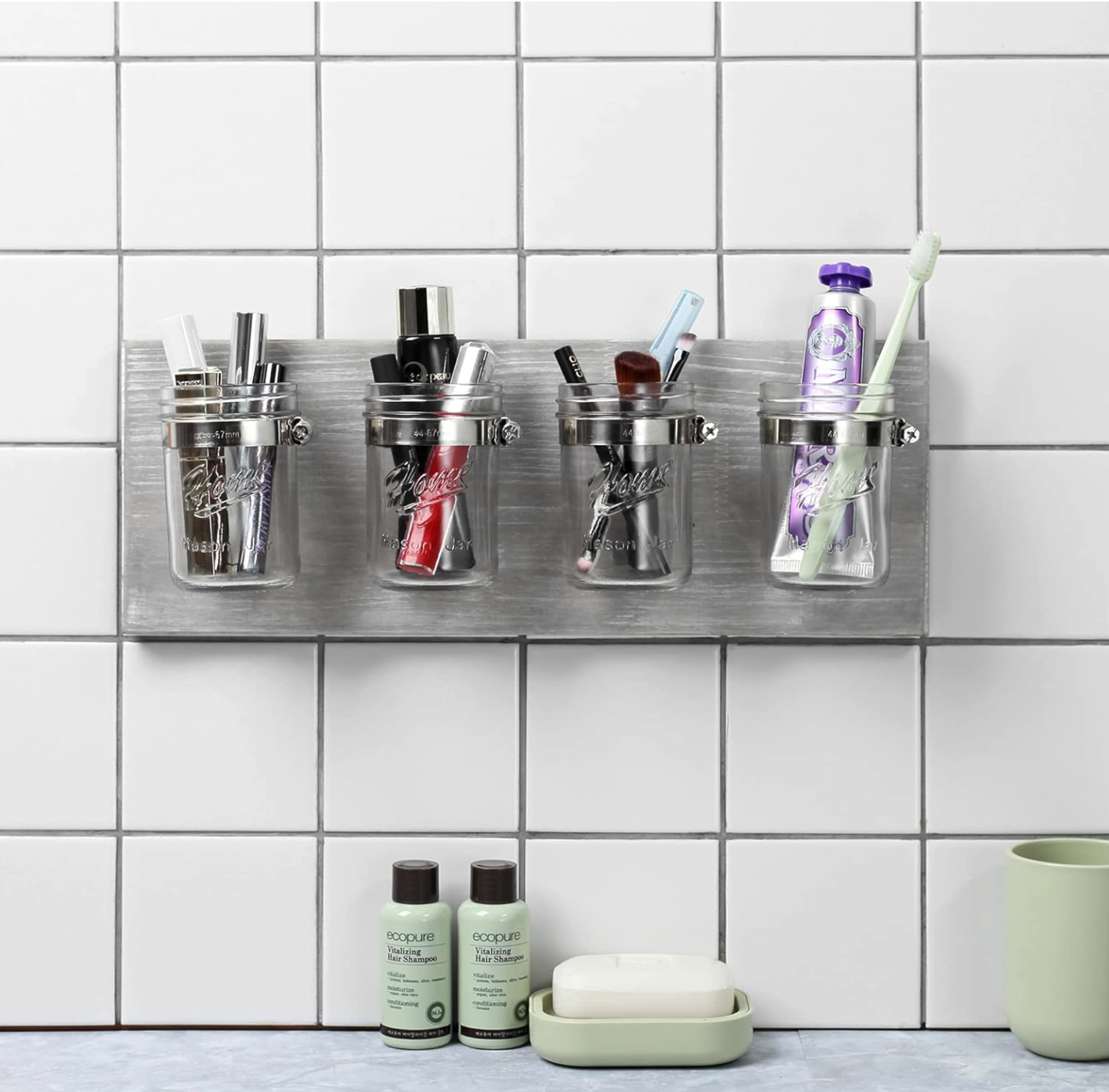 Four glass jars mounted on a tiled wall keep toiletries and toothbrushes neatly displayed above a bathroom counter with bottles and soap dishes for an organized bathroom.