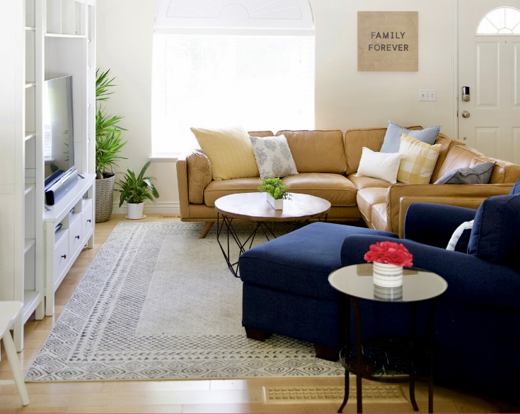 A modern living room with a tan sectional sofa, navy blue ottoman, round coffee table, and indoor plants demonstrates how to organize a small apartment. A 