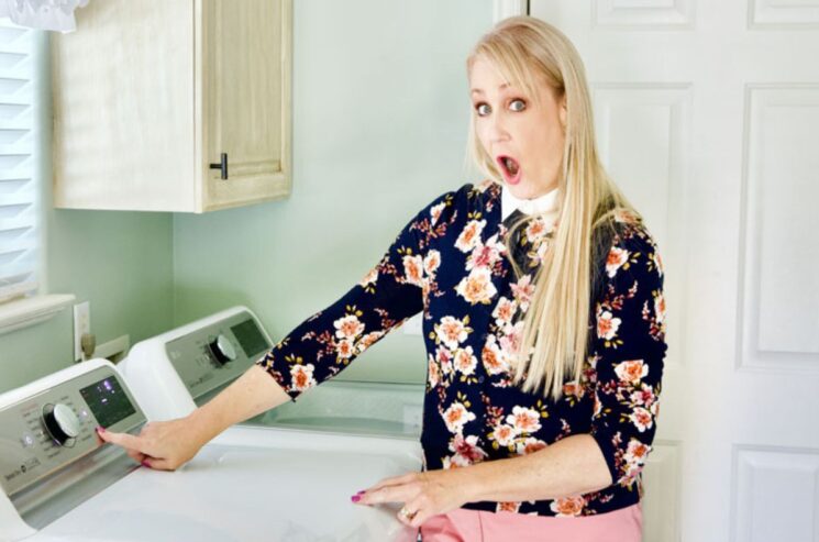 A woman with long blonde hair and a floral top presses a button on a washing machine, looking surprised—perhaps she’s just discovered the benefits of wool dryer balls for her laundry routine.