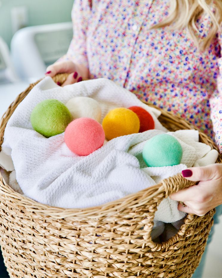 A person holds a wicker laundry basket filled with white towels and colorful wool dryer balls, highlighting the benefits of wool dryer balls for softer, fresher laundry.