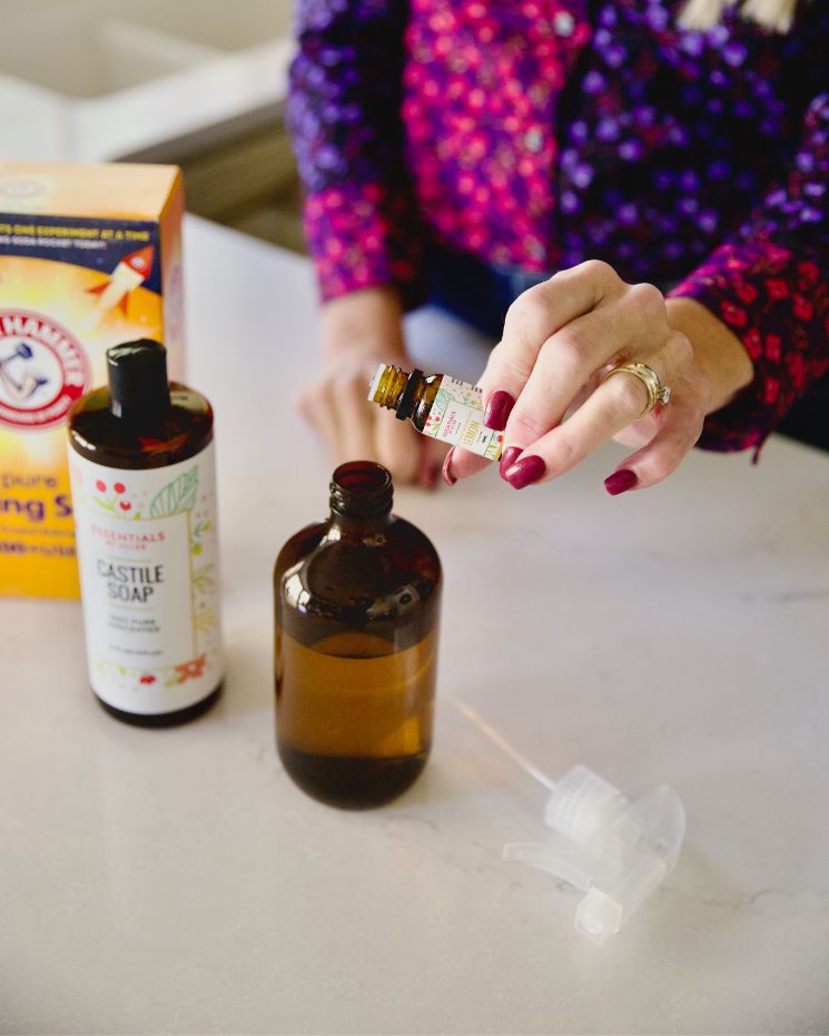 A person demonstrates castile soap uses by adding drops from an essential oil bottle into a brown glass spray bottle, surrounded by castile soap, baking soda, and a spray nozzle on a countertop.