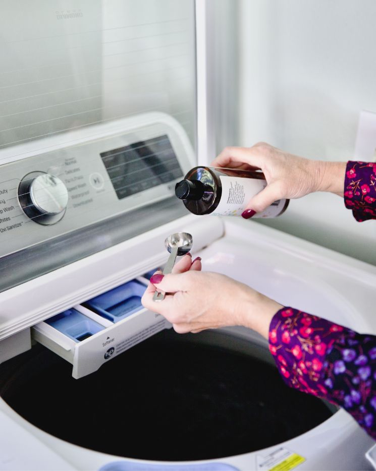 A person pours liquid laundry detergent into a measuring spoon above the detergent compartment of a top-loading washing machine, demonstrating one of the many castile soap uses for eco-friendly and effective cleaning.