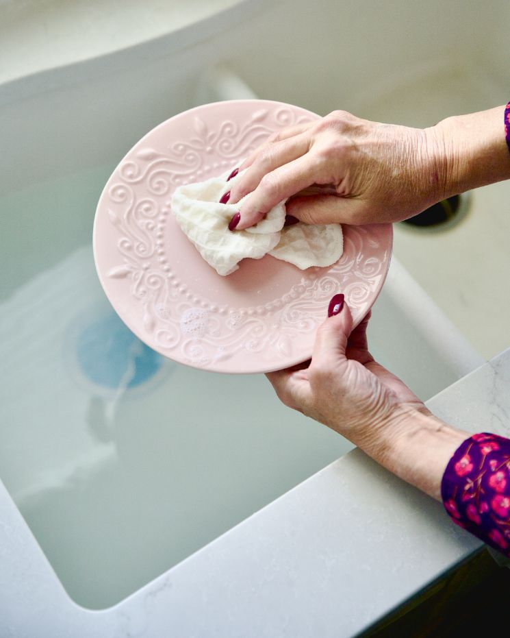 A person wipes a pink decorative plate with a white cloth over a sink filled with water, showcasing one of the many castile soap uses for gentle dish cleaning.