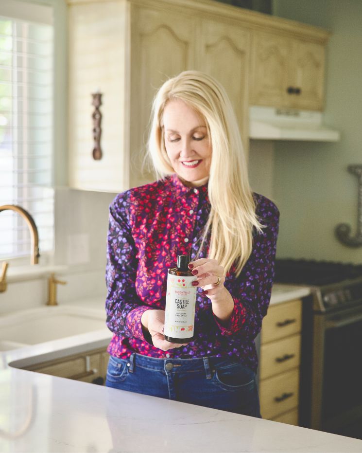 A woman with long blonde hair stands in a kitchen holding a bottle of liquid castile soap and looking at it, considering different castile soap uses for her home.