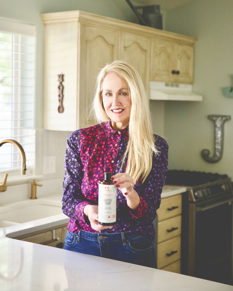 A woman stands in a kitchen holding a bottle of castile soap, smiling at the camera, ready to show off clever castile soap uses for sparkling clean counters.