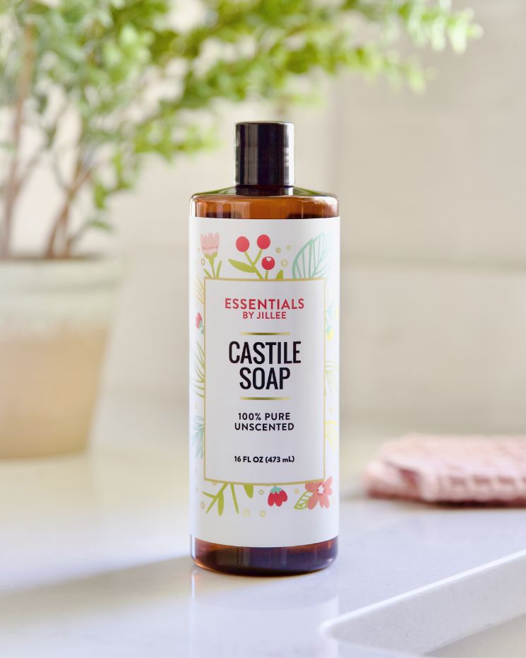 A bottle of Essentials by Jillee Castile Soap, 16 fl oz, labeled 100% pure and unscented, sits on a countertop with a blurred plant and cloth in the background—perfect for exploring versatile castile soap uses at home.