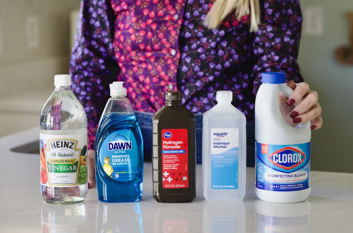 Five cleaning products—white vinegar, dish soap, hydrogen peroxide, isopropyl alcohol, and bleach—are lined up on a countertop in front of a person wearing a patterned shirt, showcasing castile soap uses alongside other staples.