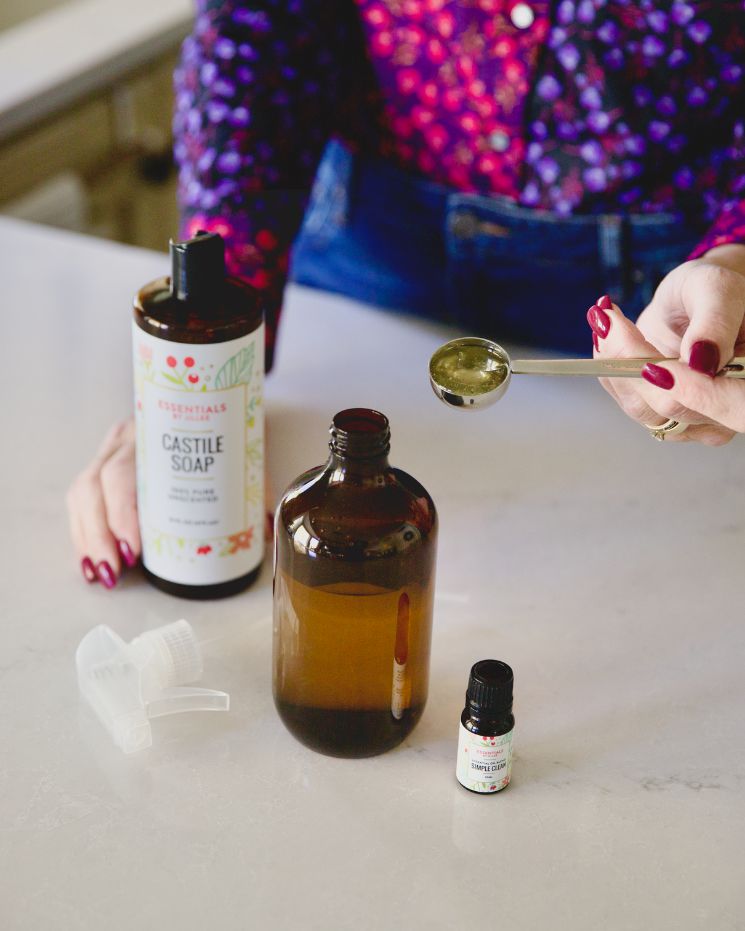 A person measures liquid with a tablespoon over an amber bottle, showing castile soap uses alongside a small essential oil bottle and spray nozzle on a white countertop.