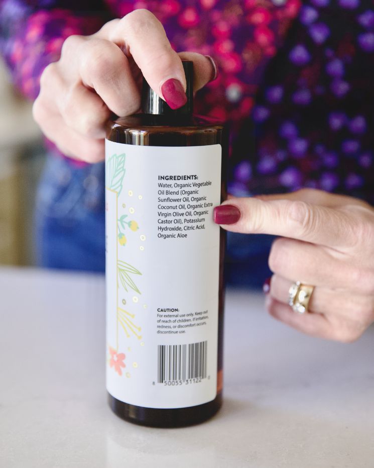 A person with painted nails points to the ingredients list on the back of a castile soap bottle with a floral label, exploring castile soap uses.