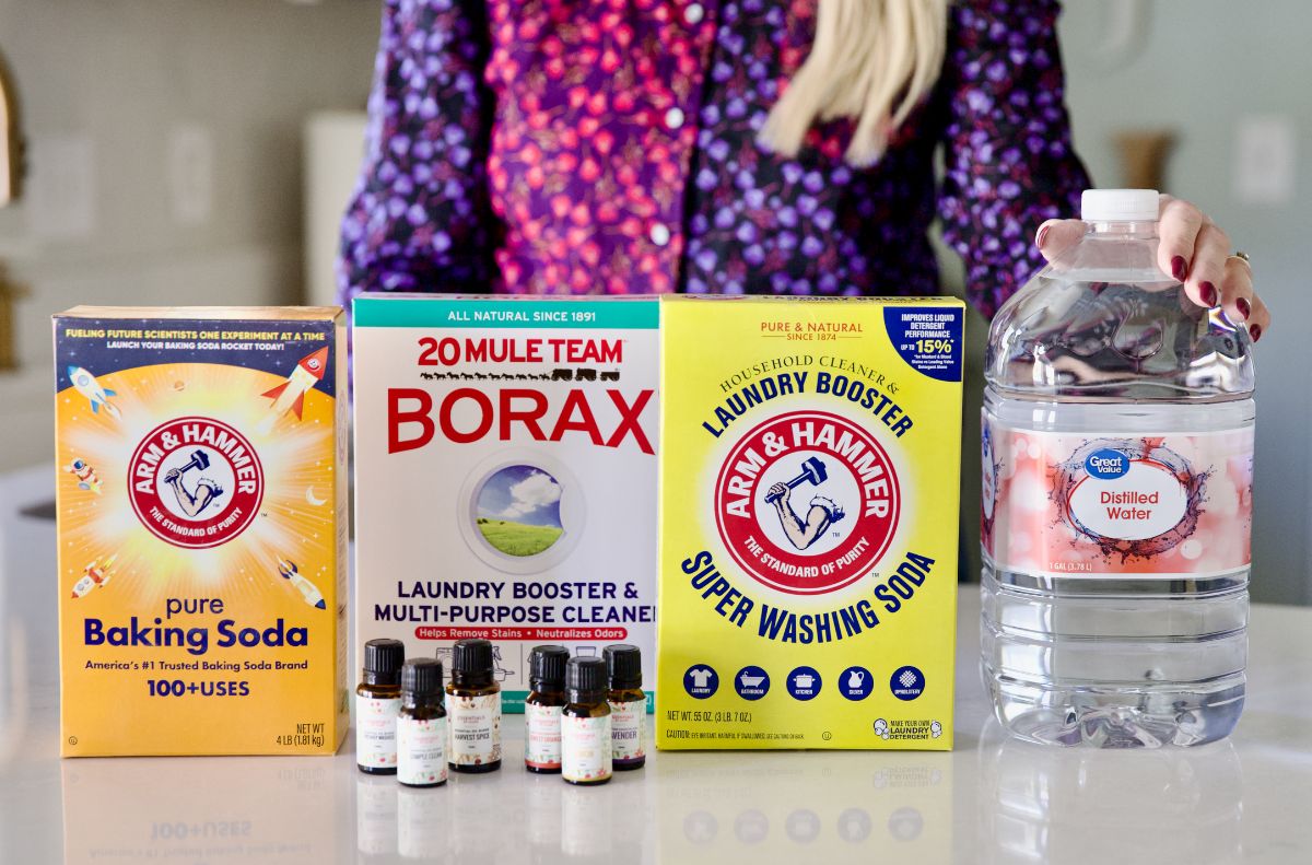 A person stands behind boxes of baking soda, Borax, washing soda, a jug of distilled water, castile soap, and several small essential oil bottles—showcasing various castile soap uses for DIY cleaning.