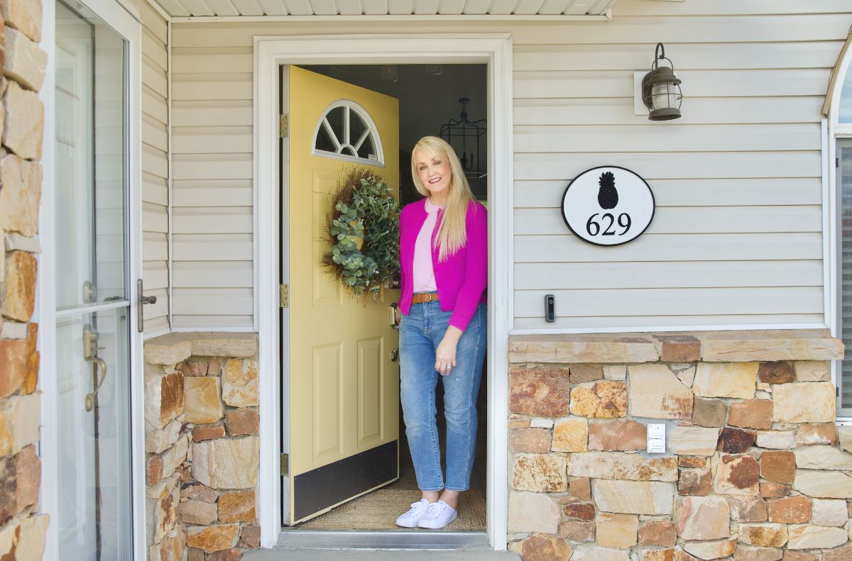 A woman stands smiling in the doorway of a house with a yellow door, stone exterior, a wreath, a calendar sign displaying the number 629, and a pineapple.