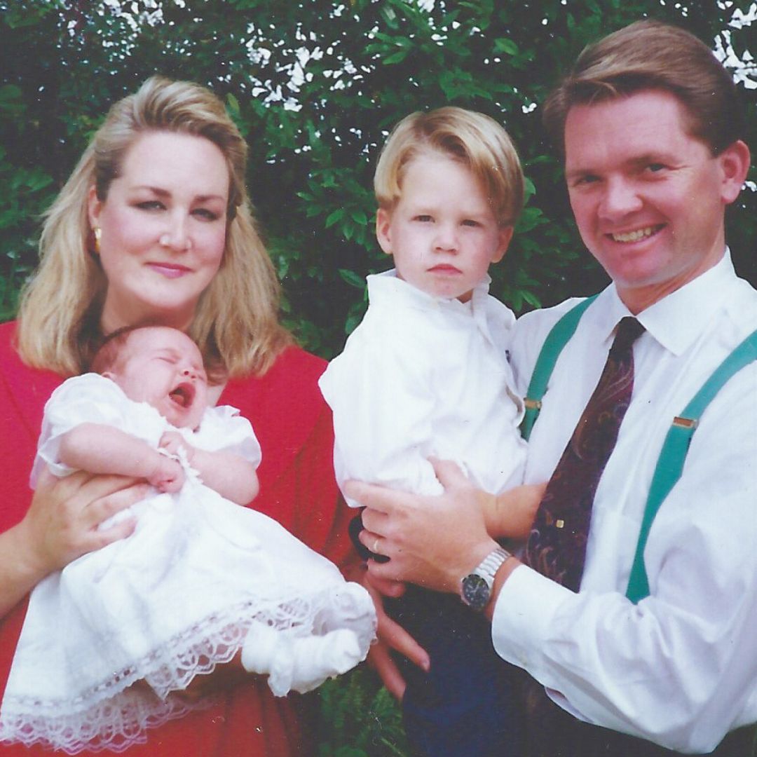 A woman holds a crying baby while a man, clutching a young boy, stands beside her. All four are dressed formally, standing outdoors in front of green foliage—perhaps posing for a family calendar photo.