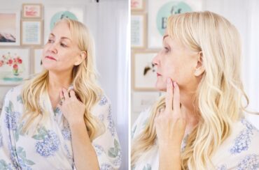 A woman with long blonde hair and a floral blouse gently touches her neck while looking in the mirror, admiring her radiant skinโperhaps thanks to vitamin Cโin both front and side profiles.