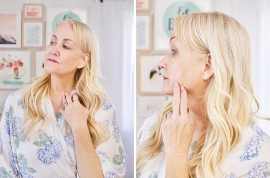 A woman with long blonde hair and a floral blouse gently touches her neck while looking in the mirror, admiring her radiant skin—perhaps thanks to vitamin C—in both front and side profiles.