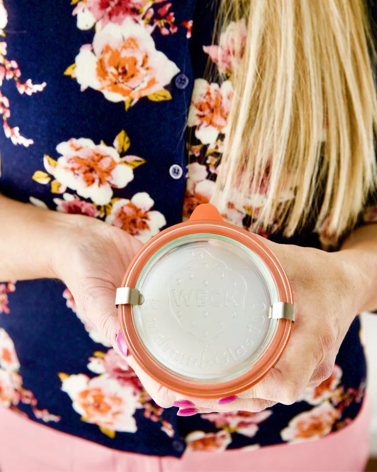 A person in a floral shirt holds a round glass container with a metal clasp and an orange rubber seal, viewed from above—perfect for storing homemade arthritis pain relief cream.