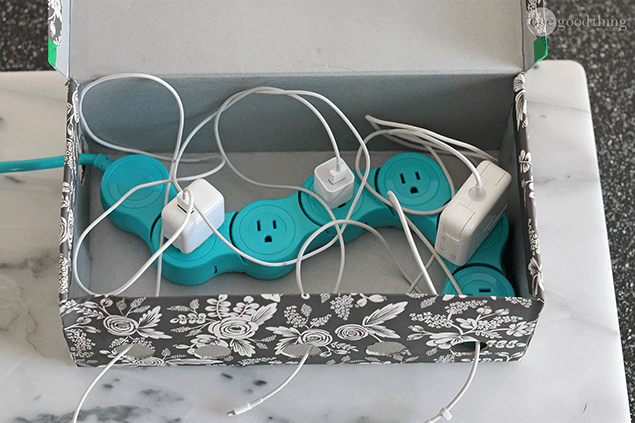 A decorative box serves as a simple charging station, housing a turquoise power strip and several tangled charging cables and plugs on a marble surface.